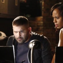 A man sits behind a desk, staring at two computer screens, while a woman stands behind him.