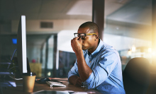 A man working at a desk puts his fingers to the bridge of his nose beneath glasses in frustration