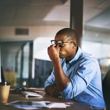 A man working at a desk puts his fingers to the bridge of his nose beneath glasses in frustration