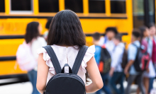 A girl wearing a backpack tentatively approaches a school bus with students waiting to board. 