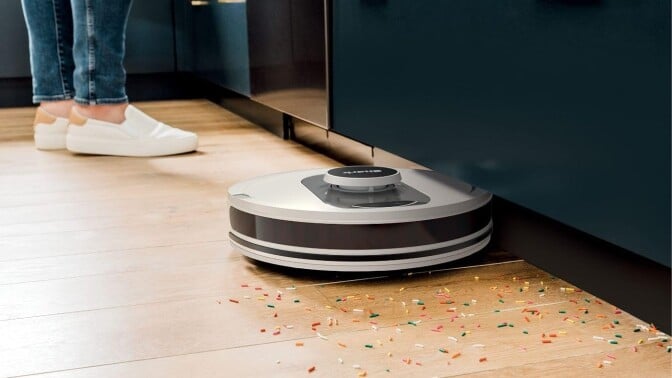 the shark ai robot vacuum cleans colorful sprinkles off a kitchen floor while someone wearing white slip-on shoes stands in the background