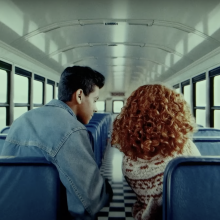 The inside of a school bus. Two students, a brown-skinned boy with a cropped hair cut and a girl with big red curly hair, are seen from the back. 