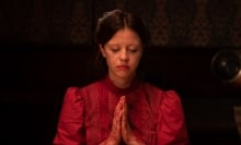 A young woman with smeary makeup and pigtails, wearing a red dress, bows her head and clasps her bloody hands in prayer.