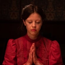 A young woman with smeary makeup and pigtails, wearing a red dress, bows her head and clasps her bloody hands in prayer.