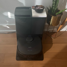 Eufy X10 Pro Omni robot vacuum and dock sitting on hardwood floor against will with potted plant and desk in peripherals