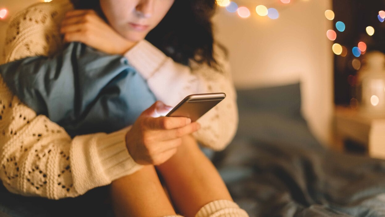 A girl dressed in a white sweater looks at her phone, while sitting in her bedroom.