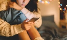 A girl dressed in a white sweater looks at her phone, while sitting in her bedroom.