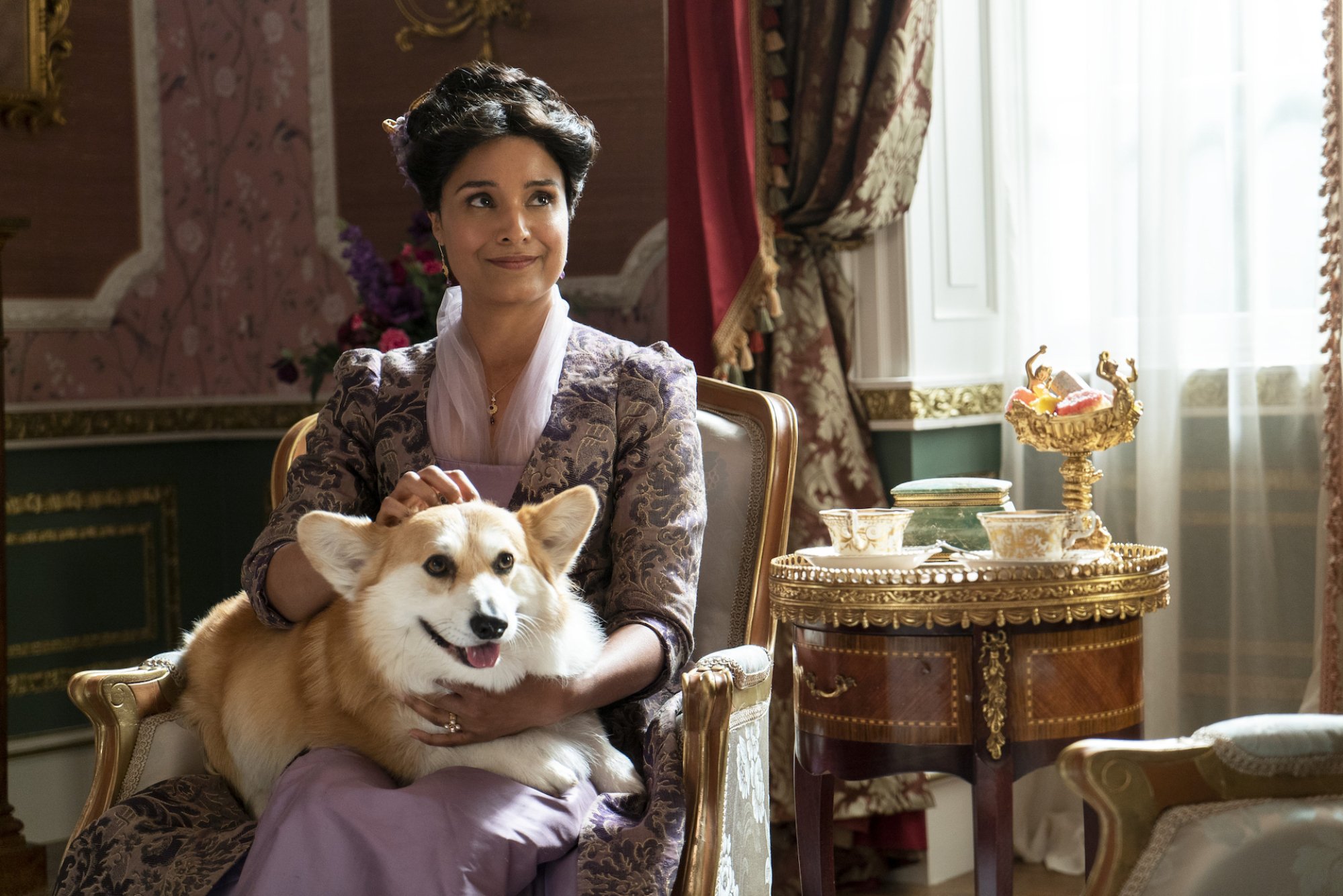 A middle aged Indian woman in Regency era clothing holding a large corgi on her lap. 