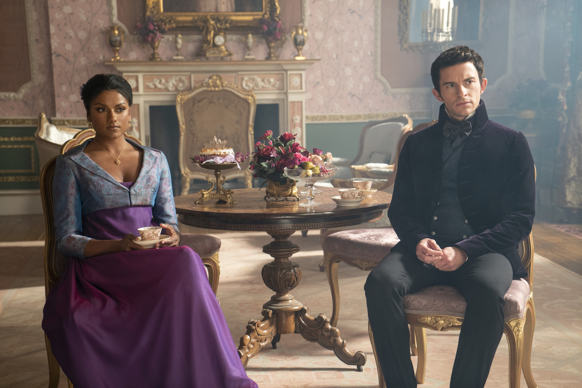 An Indian woman in a blue and purple dress sitting in a chair in a parlor. A few feet away sits a white man in a suit in another chair.