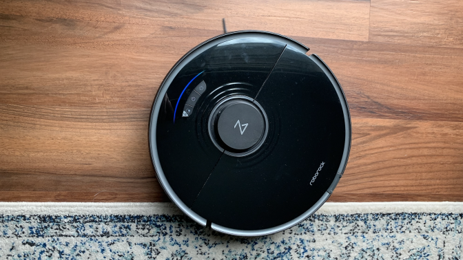 The Roborock S7, a circular, black robotic vacuum mop, on a hardwood floor next to a rug. 