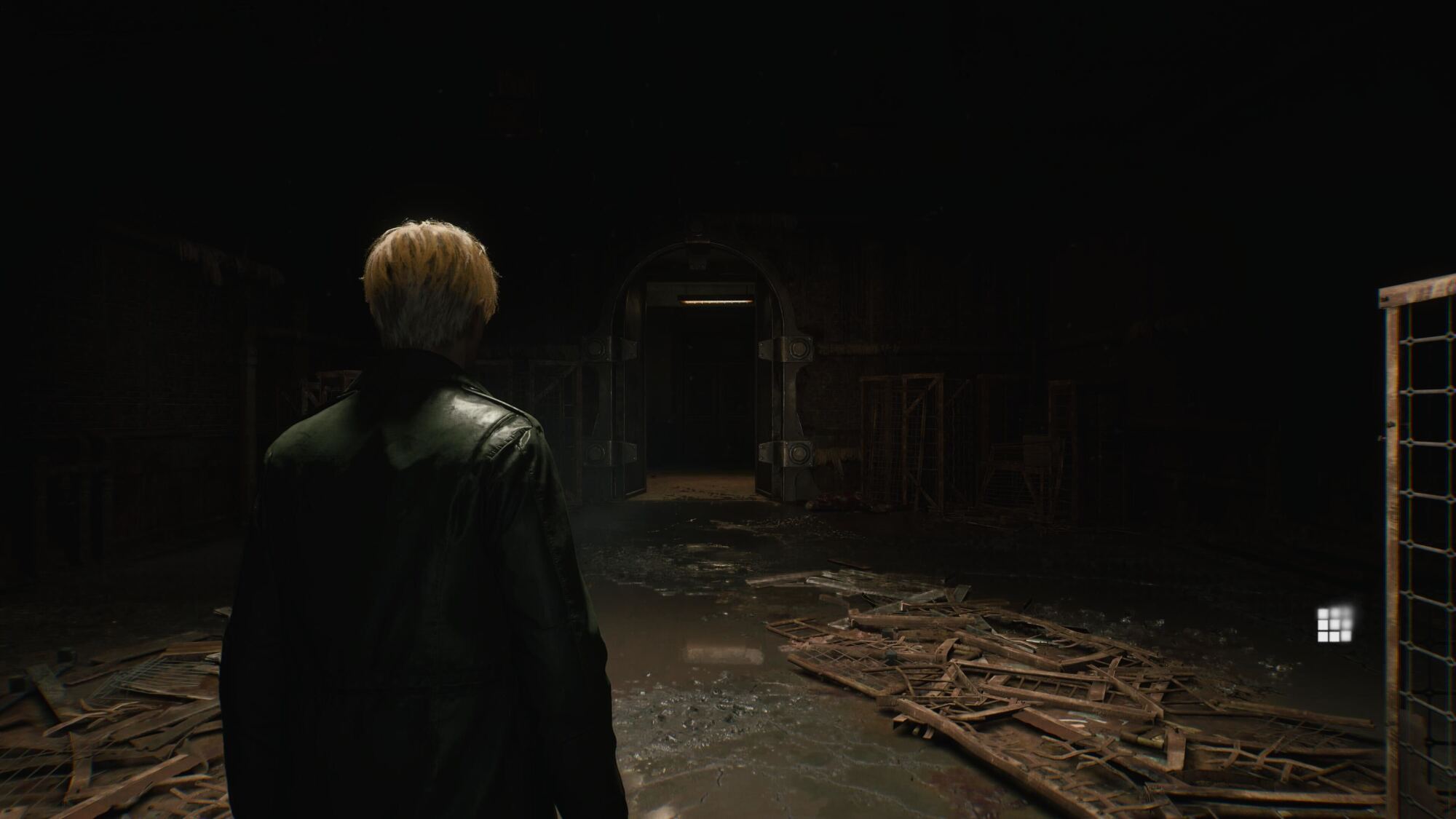 A man with light blonde hair, wearing a dark leather jacket, stands in a large, dimly lit room filled with scattered debris and broken wooden structures. He faces a large, metallic doorway that leads into a darker corridor. The scene exudes a sense of decay and abandonment, with an eerie atmosphere amplified by the minimal lighting and shadowy surroundings.