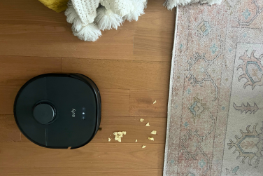 Eufy robot vacuum cleaning crumbs on hardwood floor near rug with blanket in peripheral