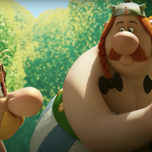 Two animated characters stand in the forest beside a boulder.