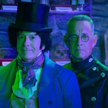 Two men in costume stand in a green lit room, staring at the camera.