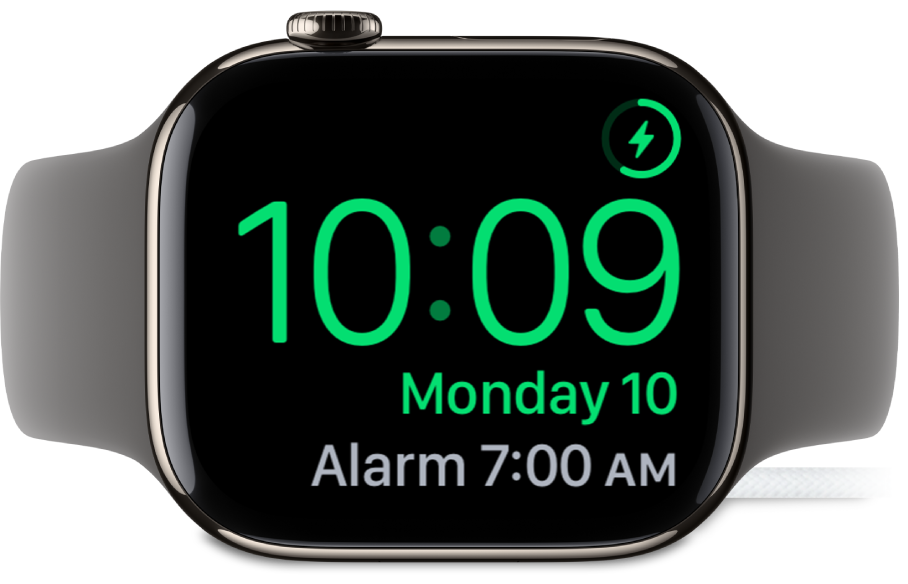 Ura Apple Watch na boku, priklopljena na polnilnik, na zaslonu pa je prikazan simbol za polnjenje v zgornjem desnem kotu, trenutni čas pod njim in čas naslednjega alarma.