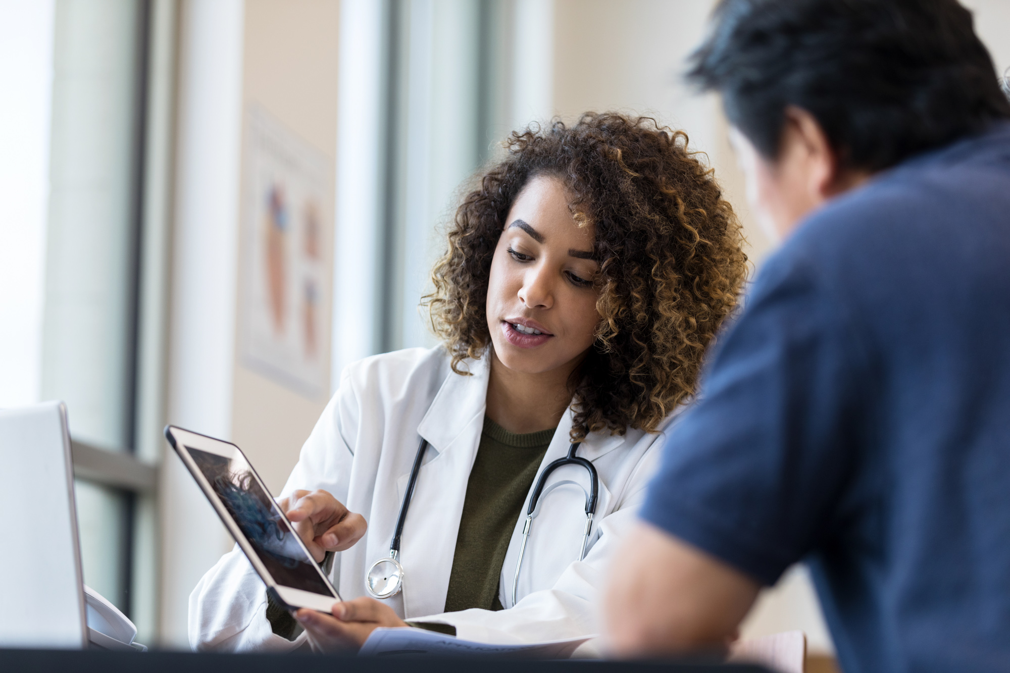 Young adult female doctor uses the digital tablet to show data to the unrecognizable senior man