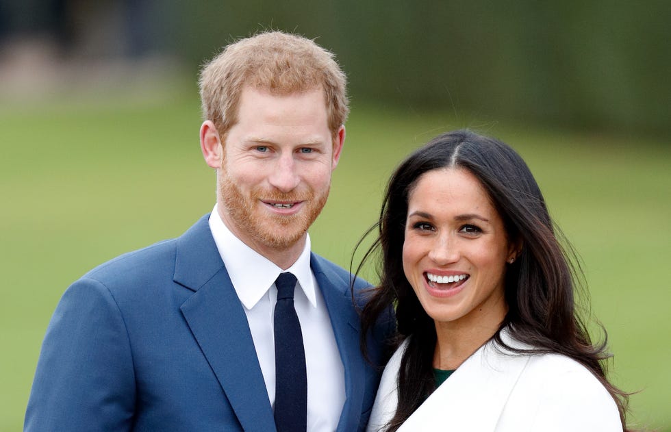 prince harry and meghan markle smile and stand together outside, he wears a blue suit jacket, white collared shirt and navy tie, she wears a white coat over a dark top