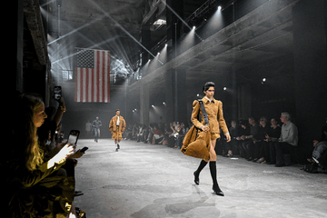 a woman in a tan outfit and oversized bag walking down a runway with an american flag in the background