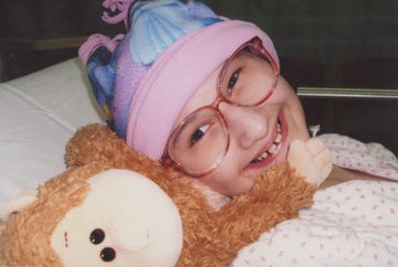 a girl wearing glasses and a pink and purple hat smiles as she lies in a bed next to a stuffed animal