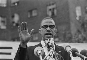 malcolm x speaks into several microphones, he has his right hand raised in front of him, he wears a suit and his signature glasses