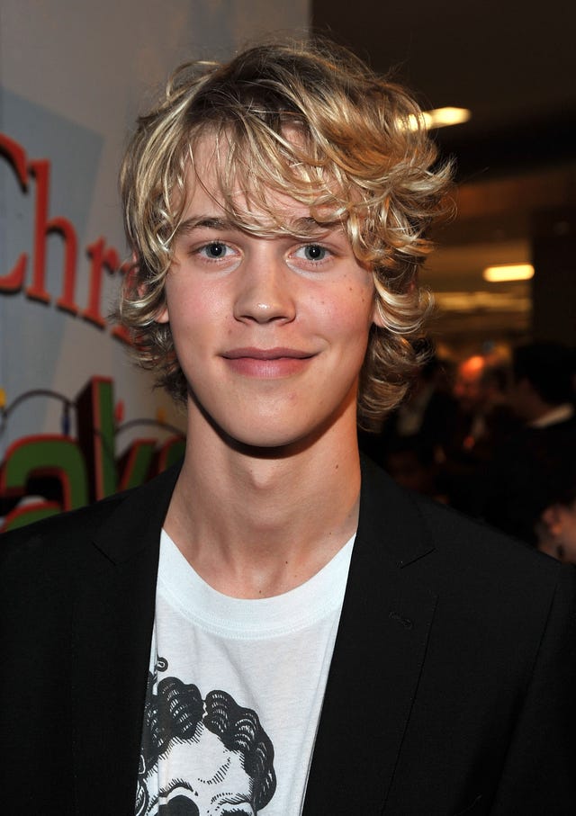 austin butler smiles at the camera, he wears a black cardigan over a white graphic tshirt