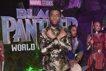 chadwick boseman stands with his arms crossed in an x over his chest, he is in front of a black panter movie poster and wears a black and gold shirt