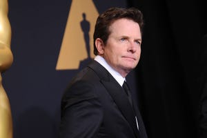 michael j fox stands on the red carpet at the oscars, he is wearing a black suit and tie with a white collared shirt and stands in front of a backdrop with an oscars logo on it, he looks past the camera with a neutral expression on his face