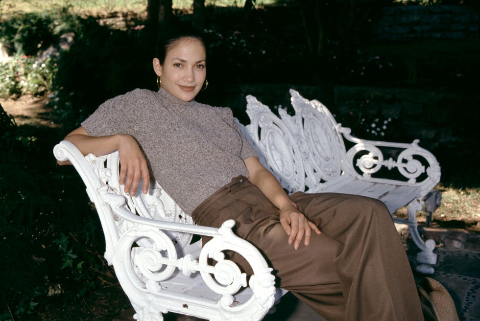 jennifer lopez smiles at the camera while sitting on a bench outside, she wears a short sleeve blouse and brown pants