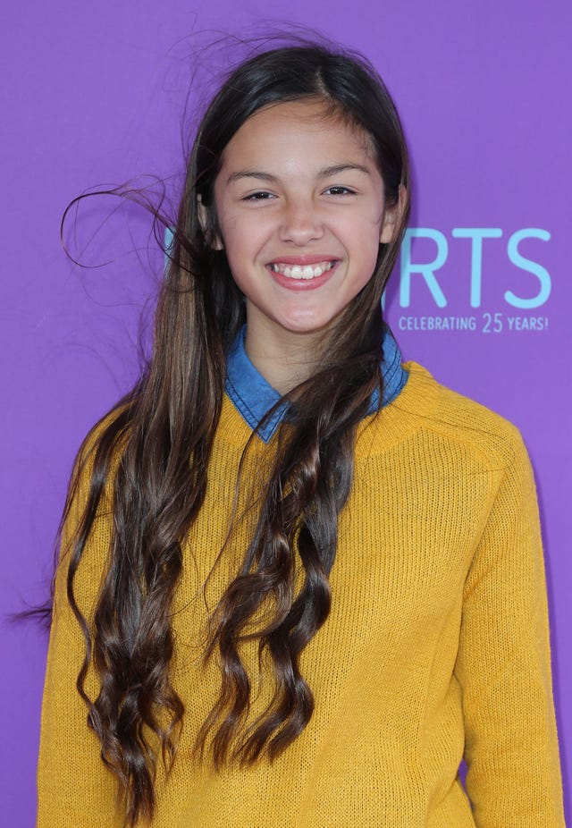 olivia rodrigo smiles for a photo in a golden yellow sweater over a blue collared shirt