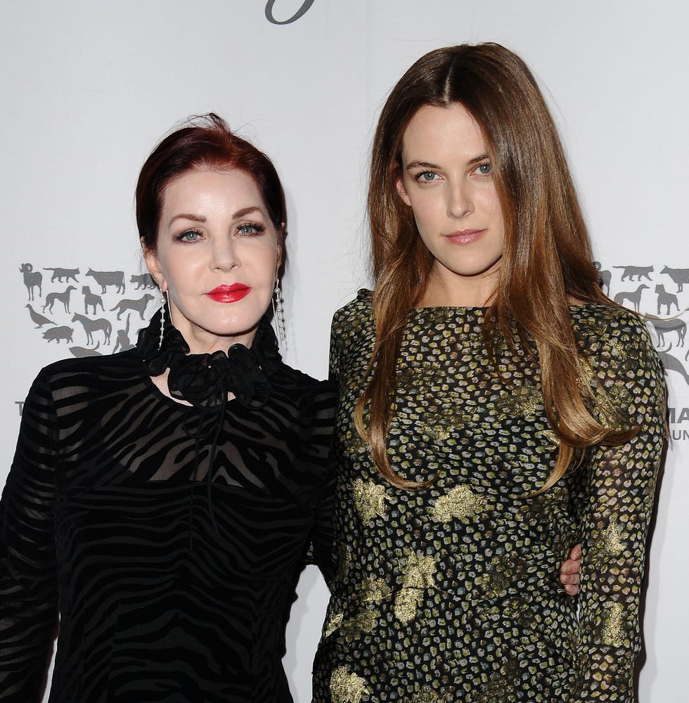 priscilla presley and riley keough pose for a photo together and smile at the camera, presley wears a black sheer dress, keough wears a green and black patterned long sleeve dress