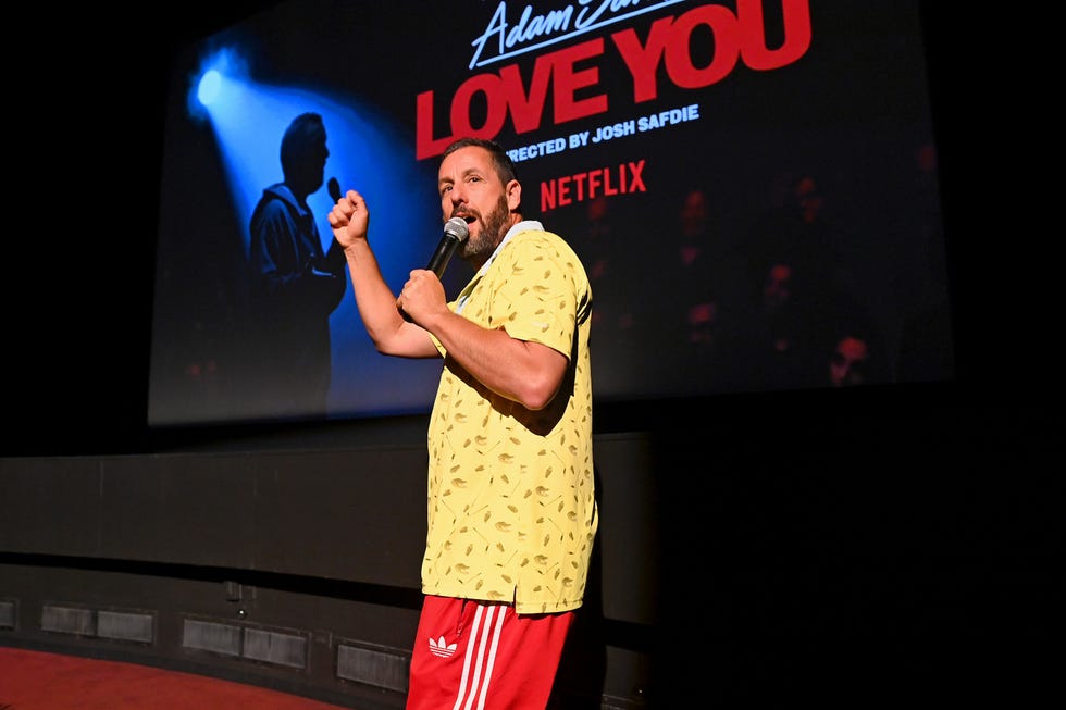 adam sandler talks into a microphone he holds in one hand while standing in front of a screen that projects his comedy special love you