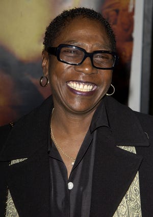 afeni shakure smiles and looks off camera, she is wearing black glasses, hoop earrings and a black outfit