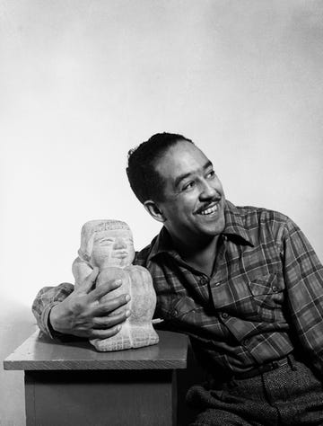 langston hughes smiles and looks right while leaning against a desk and holding a statue sitting on it, he wears a plaid shirt and pants