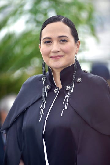 lily gladstone wearing a black outfit and native american jewelry, smiling directly at the camera