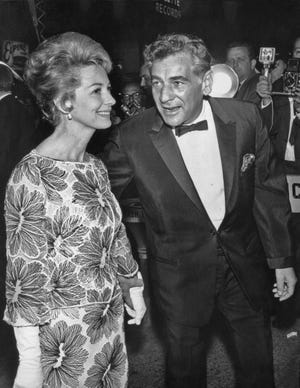 a black and white photo of felicia montealegre, wearing a floral dress, and leonardo bernstein, wearing a tuxedo, walking past reporters and photographers