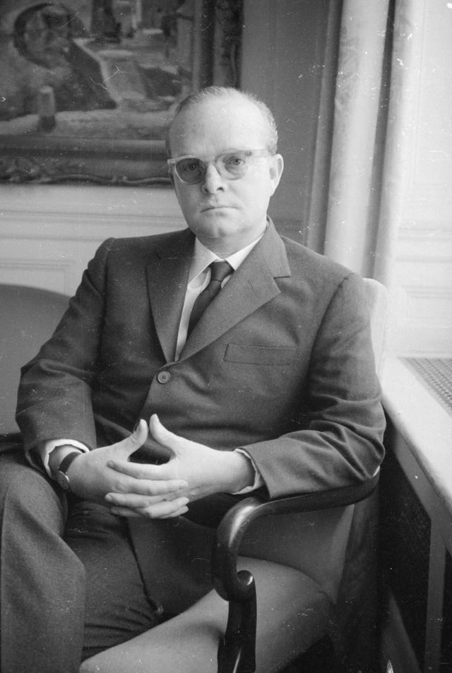 truman capote sits in an armchair and looks at the camera, he wears a suit with a tie, glasses, and a watch, his hands are clasped on his lap