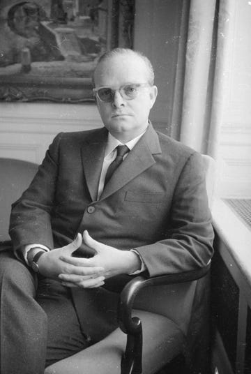 truman capote sits in an armchair and looks at the camera, he wears a suit with a tie, glasses, and a watch, his hands are clasped on his lap