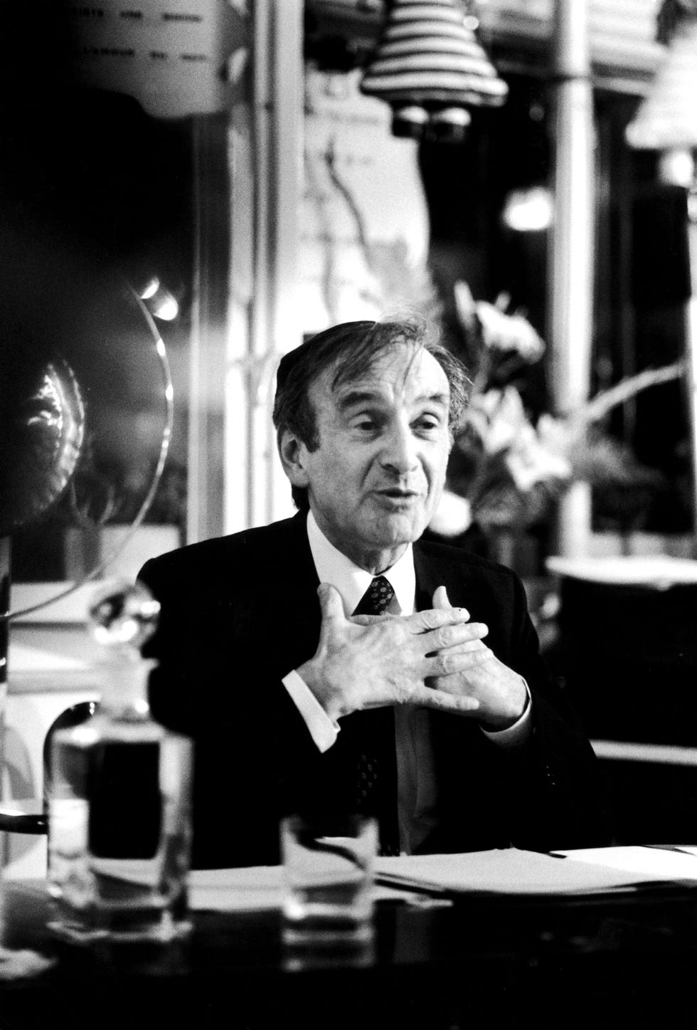 elie wiesel talks as he sits at a table in a suit jacket, dress shirt and tie with his hands in front of his chest