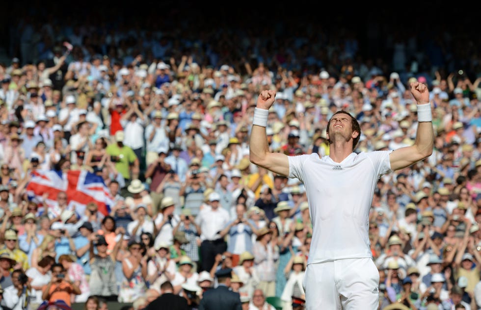 andy murray raises both arms up and lifts his head in victory, behind him a large crowd sits in the stands, murray wears an all white athletic outfit and wrist bands