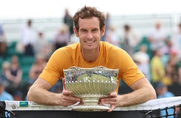 andy murray smiles at the camera while holding a silver bowl trophy, he wears an orange t shirt and leans against a tennis net