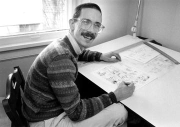 bill watterson, wearing glasses and a mustache, smiles at the camera while sitting at a desk and drawing a cartoon