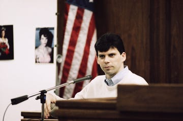 lyle menendez sits on the witness stand in a wood paneled room, he wears a white sweater and a blue collared shirt, behind him is an american flag and a white bulletin board with a blurry photo of a woman