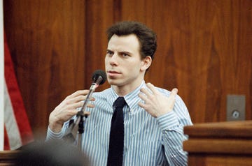 erik menendez talks into a microphone while seated in a wooden paneled room, he wears a blue and white striped collared shirt with a black tie and holds his hands in front of his chest