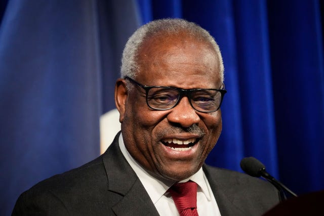 supreme court justice clarence thomas smiling and standing in front of a microphone, he is wearing a black suit jacket, white collared shirt, red tie, and black framed glasses