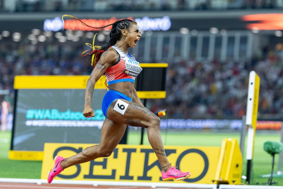 sha'carri richardson is seen in profile running on a track in celebration, she screams in triumph and wears a red white and blue usa uniform with a number 6 on her leg and pink shoes, a stadium with a crowd is in the background