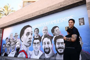 brittney griner hugs someone on her left and looks over her shoulder, she is smiling and both people wear all black outfits, they stand next to a painted mural of 15 people and the slogan bring our families home