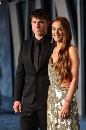 ben smith petersen and riley keough stand together and pose for a photo, he wears an all black suit and tie, she wears a glittering gold dress and smiles at the camera