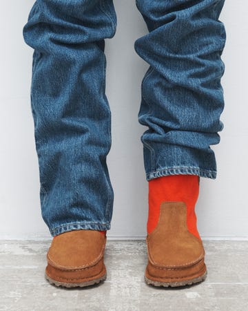 the image showcases a pair of feet wearing two different types of footwear one foot features a brown suede slipon shoe while the other is adorned with an orange sock protruding from a similar style of shoecreating a striking color contrast the individual is dressed in blue denim jeans that are slightly rolled at the ankles the background is a plain white wall and the floor appears to be a lightcolored textured surfac