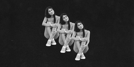 three identical figures seated on the ground each with their legs crossed and arms resting on their knees the figures have long hair and wear casual clothing including a fitted top and sneakers the background is uniformly black enhancing the focus on the figures