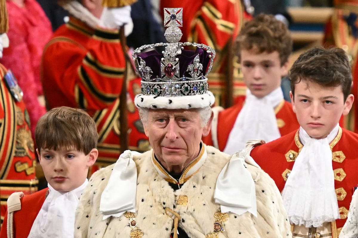 king charles walks down on an aisle with three boys following, he smiles slightly and wears a purple and bejeweled crown with a cream fur robe